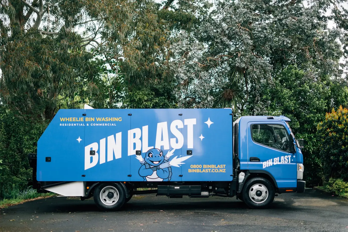 Side view of the custom blue Bin Blast wheelie bin washing truck parked under trees, with the turtle mascot on the body.
