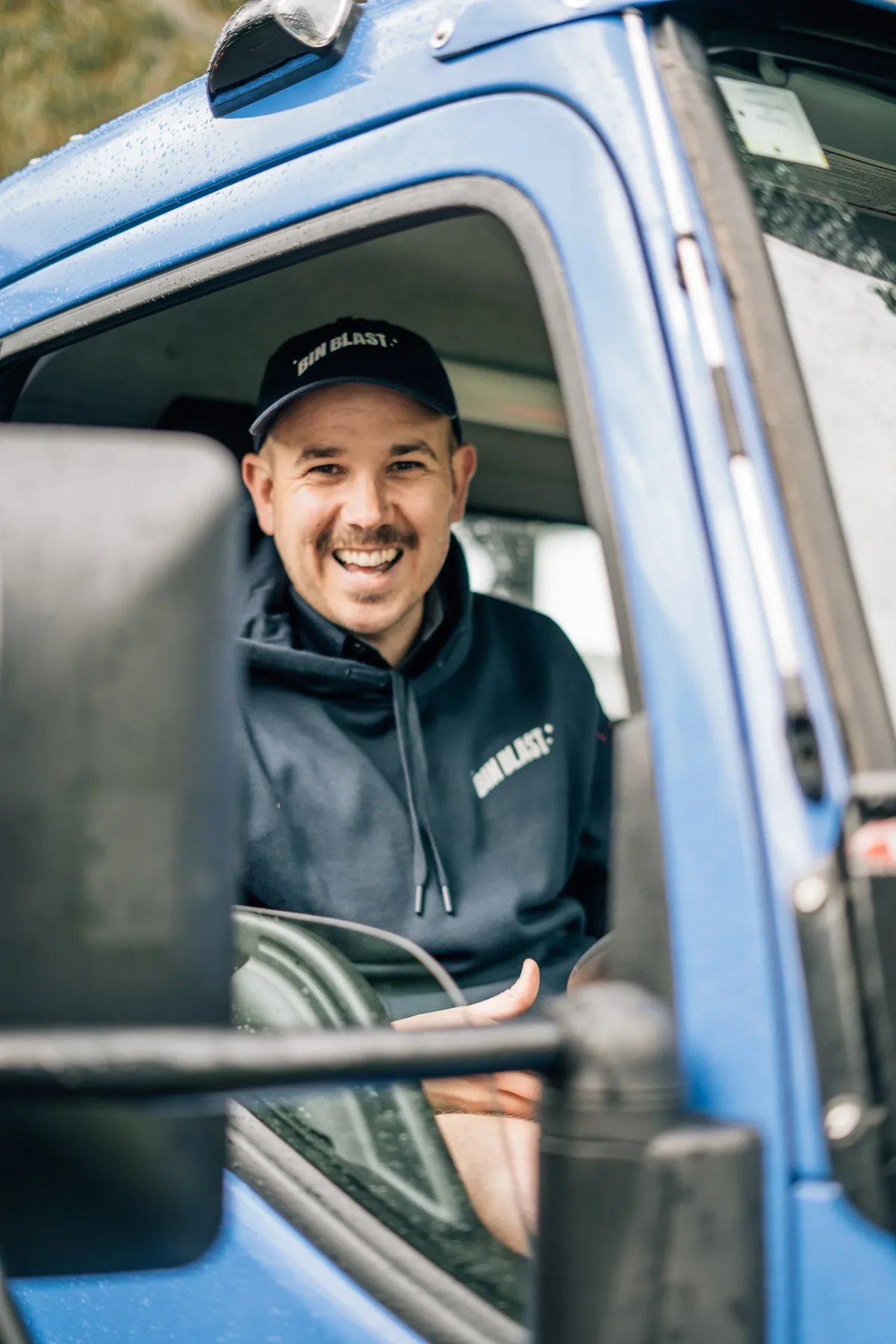 Smiling Bin Blast team member in a cap and hoodie leaning out of the blue truck cab window.